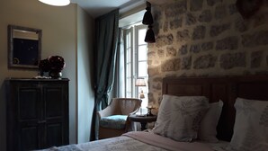 A bedroom with a stone wall, a bed with floral bedding, a chair, and a small table with a lamp. - Chateau Le Colombier - Chambres d'hôtes (Carentan les Marais)