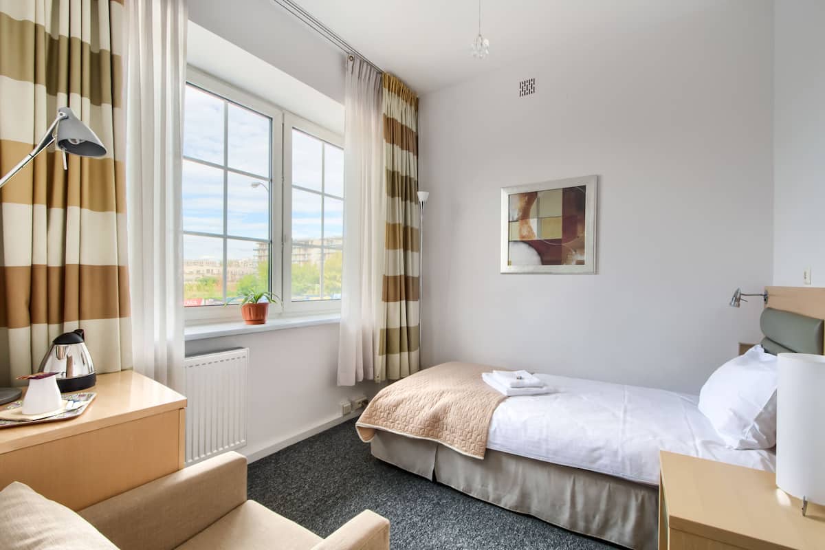 Een hotelkamer met een bed, bureau, stoel, raam met gordijnen, radiator, een plant en een schilderij aan de muur.