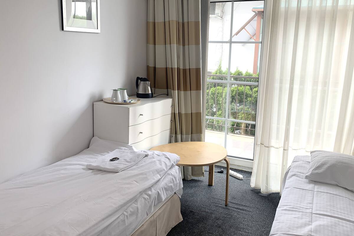 Een kleine, schone kamer met twee enkele bedden, een bureau, een stoel, een koffiezetapparaat en een raam met gordijnen.
