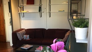 Una sala de estar con un sofá marrón oscuro, una mesa de centro con revistas, una planta en maceta y una estantería con libros y adornos.
