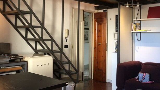 A room with a staircase, a white appliance labeled 'SMEG', a red couch, and a bookshelf.