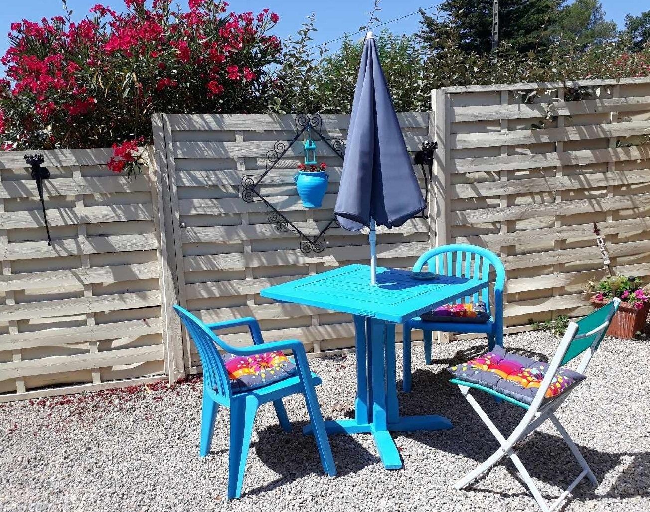 Et lille udendørs siddeområde med et bord og to stole, en blå parasol og en hængende blå plantekasse.