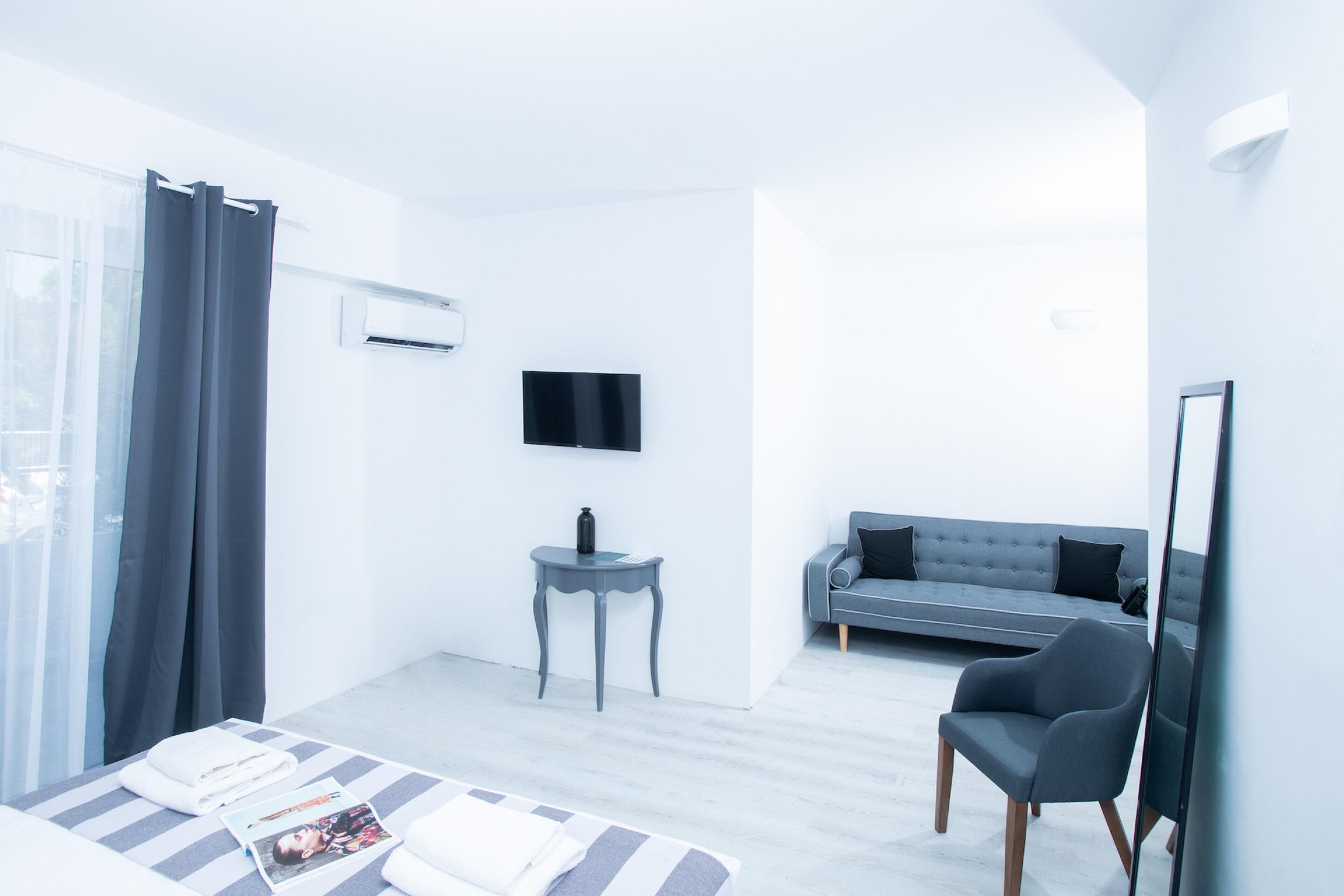 Ein modernes Hotelzimmer mit einem Bett, einem grauen Sofa, einem kleinen Beistelltisch, einem an der Wand montierten Fernseher und einem großen Spiegel.