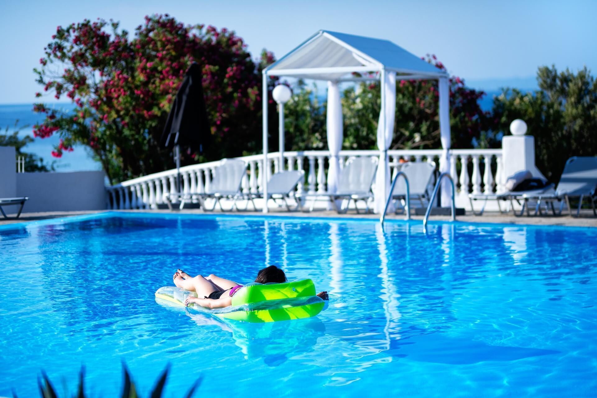 A pool with a person on a green float, lounge chairs, and a gazebo in the background.
