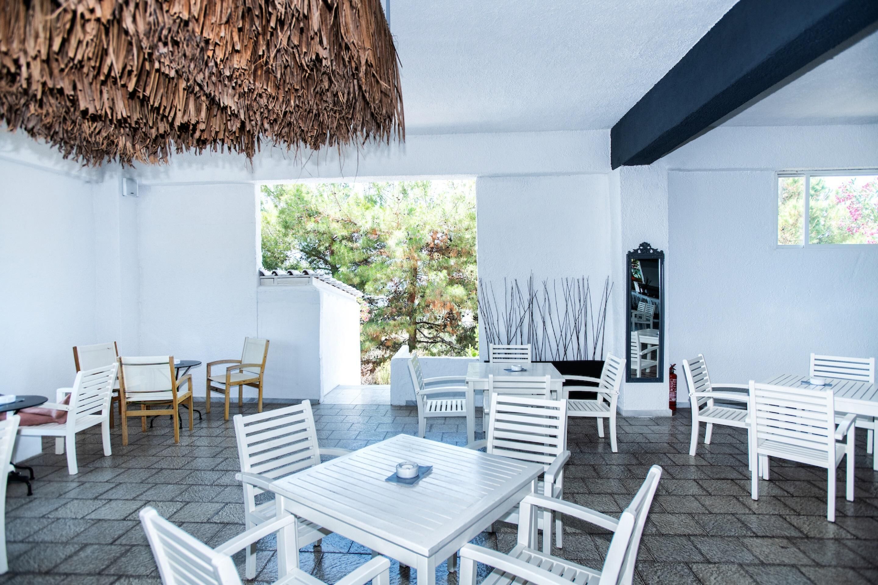 Un comedor moderno con mobiliario blanco, techo de paja y grandes ventanas que dan a un paisaje de árboles.