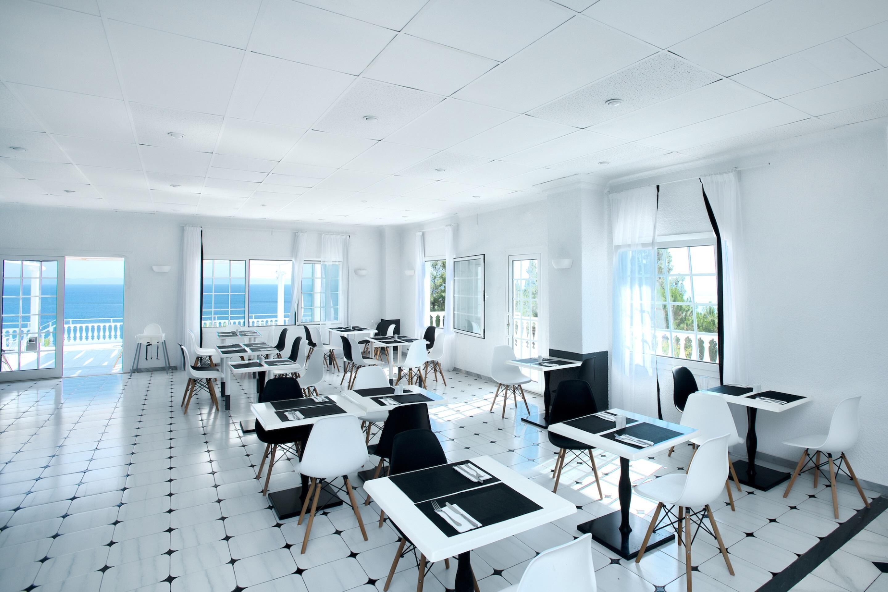 A spacious room with multiple tables and chairs, large windows with a view of the sea, and a tiled floor.