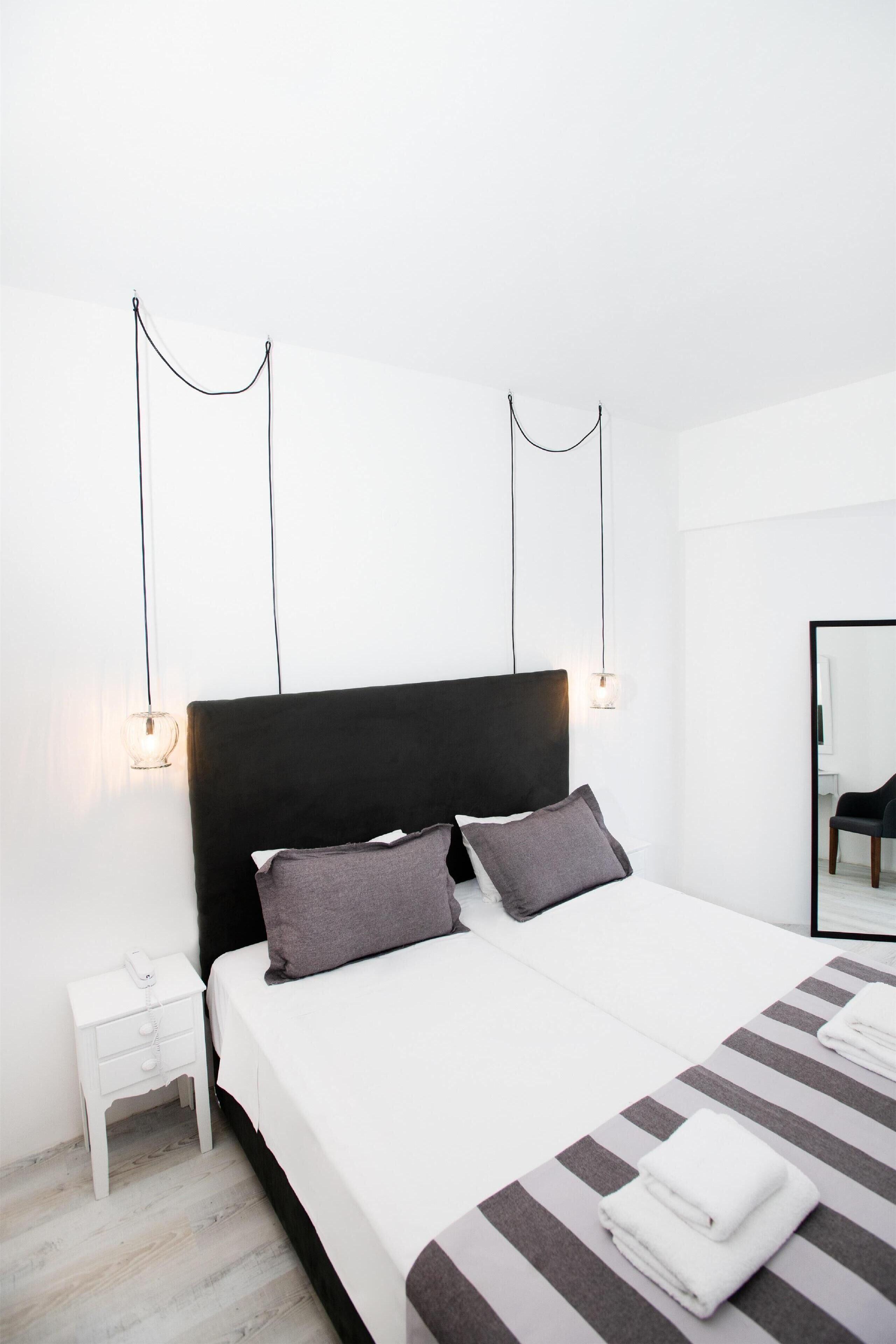Une chambre d’hôtel moderne avec un grand lit, des murs blancs et une tête de lit noire. On y trouve deux tables de chevet avec des lampes, un miroir au mur et un tapis à rayures.