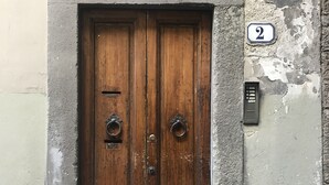 A wooden double door with a metal grid window above, a number '2' sign, and a small ventilation unit on the wall. - Vecchia Volta (Florence)