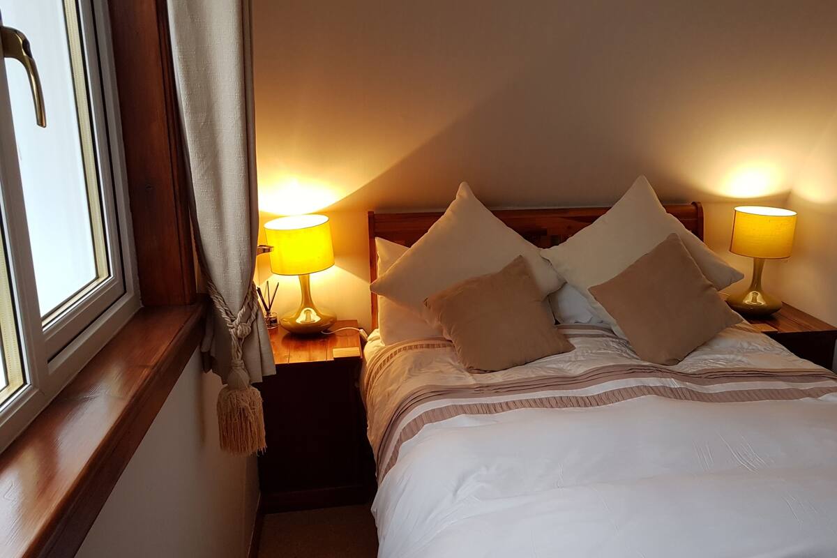 A neatly made bed with white linens and multiple pillows, two bedside lamps, and a window with curtains.