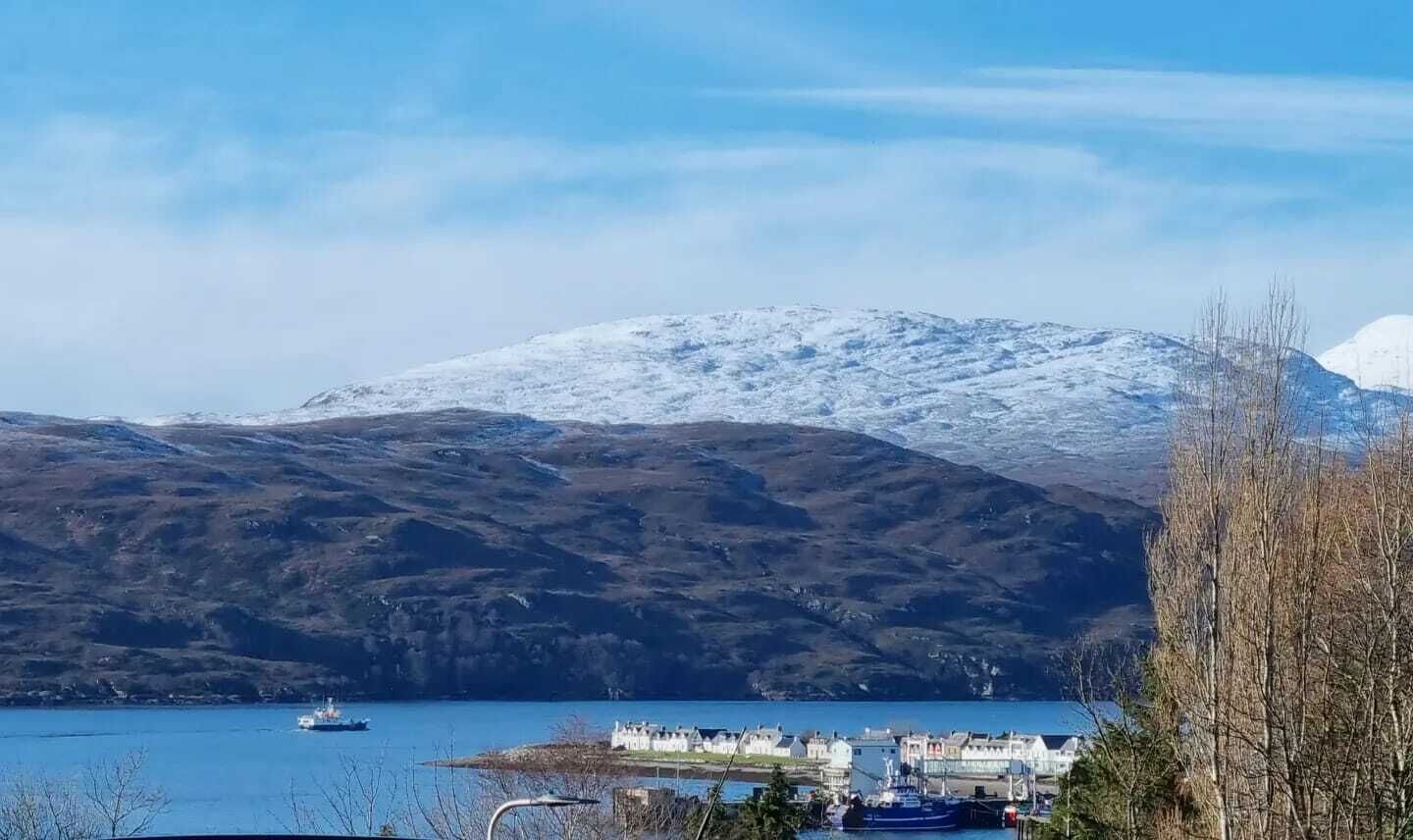 A coastal town with a snow-covered mountain in the background, a small harbor with boats, and a clear blue sky.