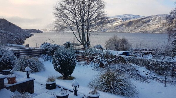 A snowy landscape with a lake, a bench, and a tree in the foreground.