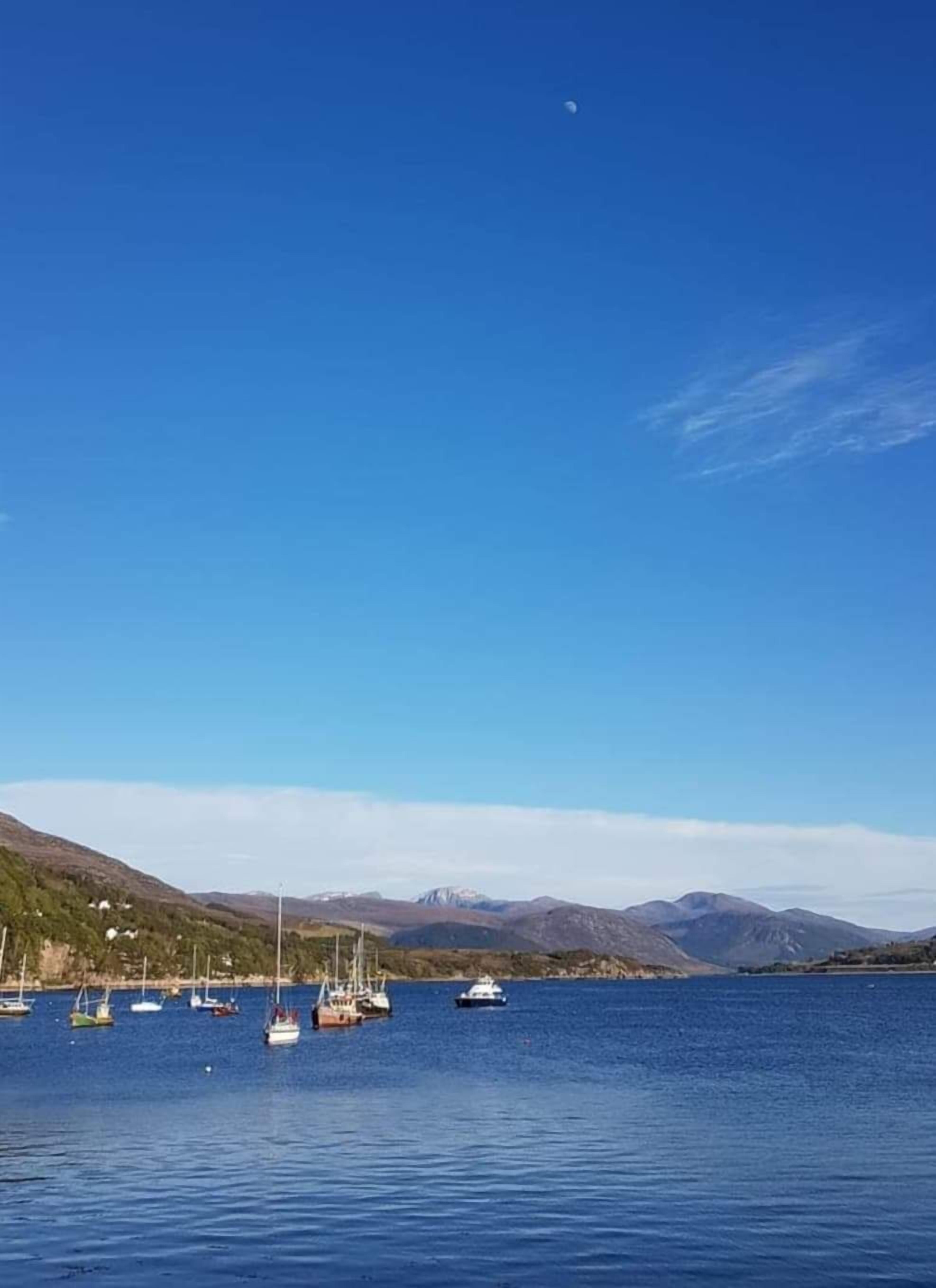 A calm lake with several boats anchored, surrounded by hills and clear skies.