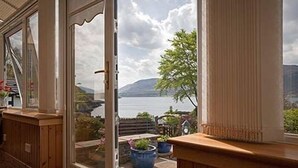 A room with a large window overlooking a lake and mountains, wooden furniture, and potted plants. - Lochview Guest House (Ullapool)