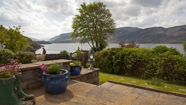 A stone patio with potted plants, a tree, and a view of a lake and mountains. - Lochview Guest House (Ullapool)