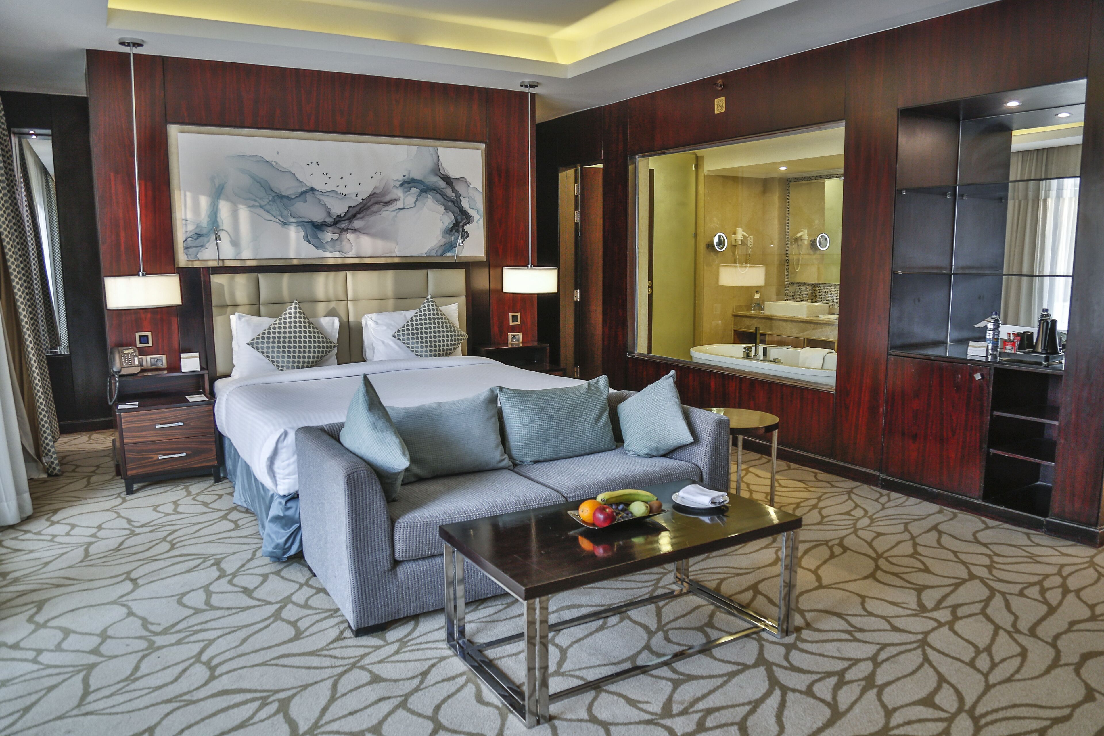 A hotel room with a large bed, a sofa, a small table with fruit, a bathroom with a mirror, and a large artwork on the wall.
