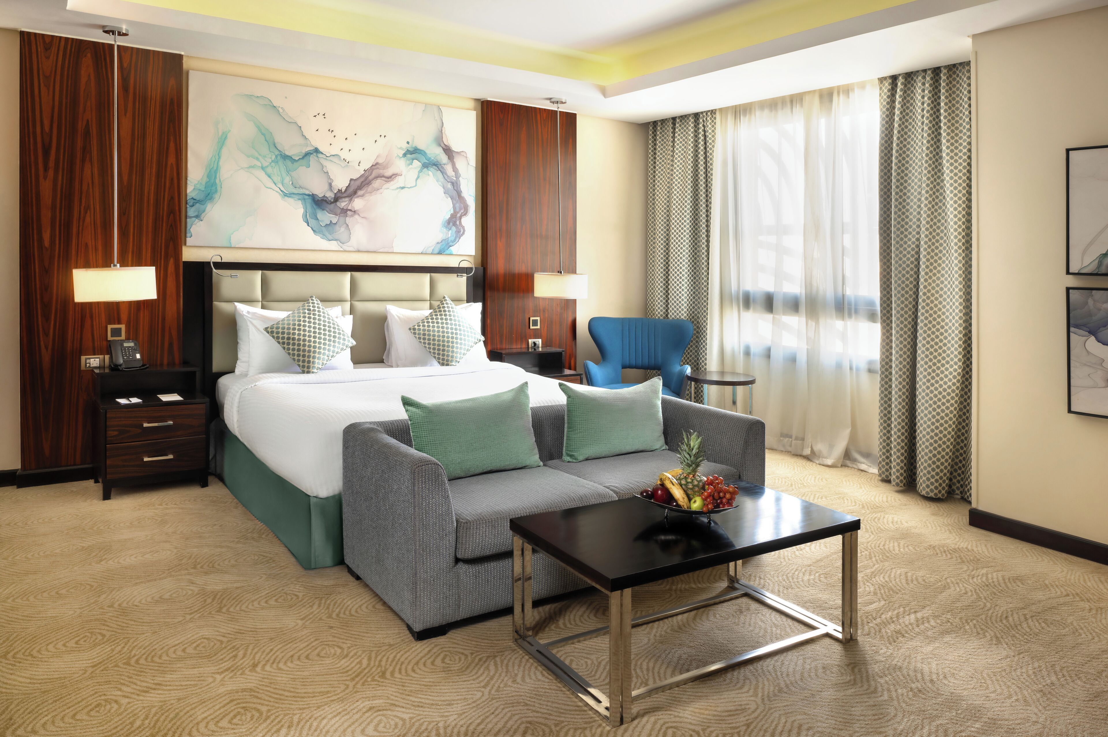 Quarto de hotel com uma cama grande, um sofá cinza, uma pequena mesa com frutas e uma cadeira azul.