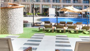 A poolside area with lounge chairs, a fruit bowl, and a view of a hotel building with multiple balconies and a swimming pool.