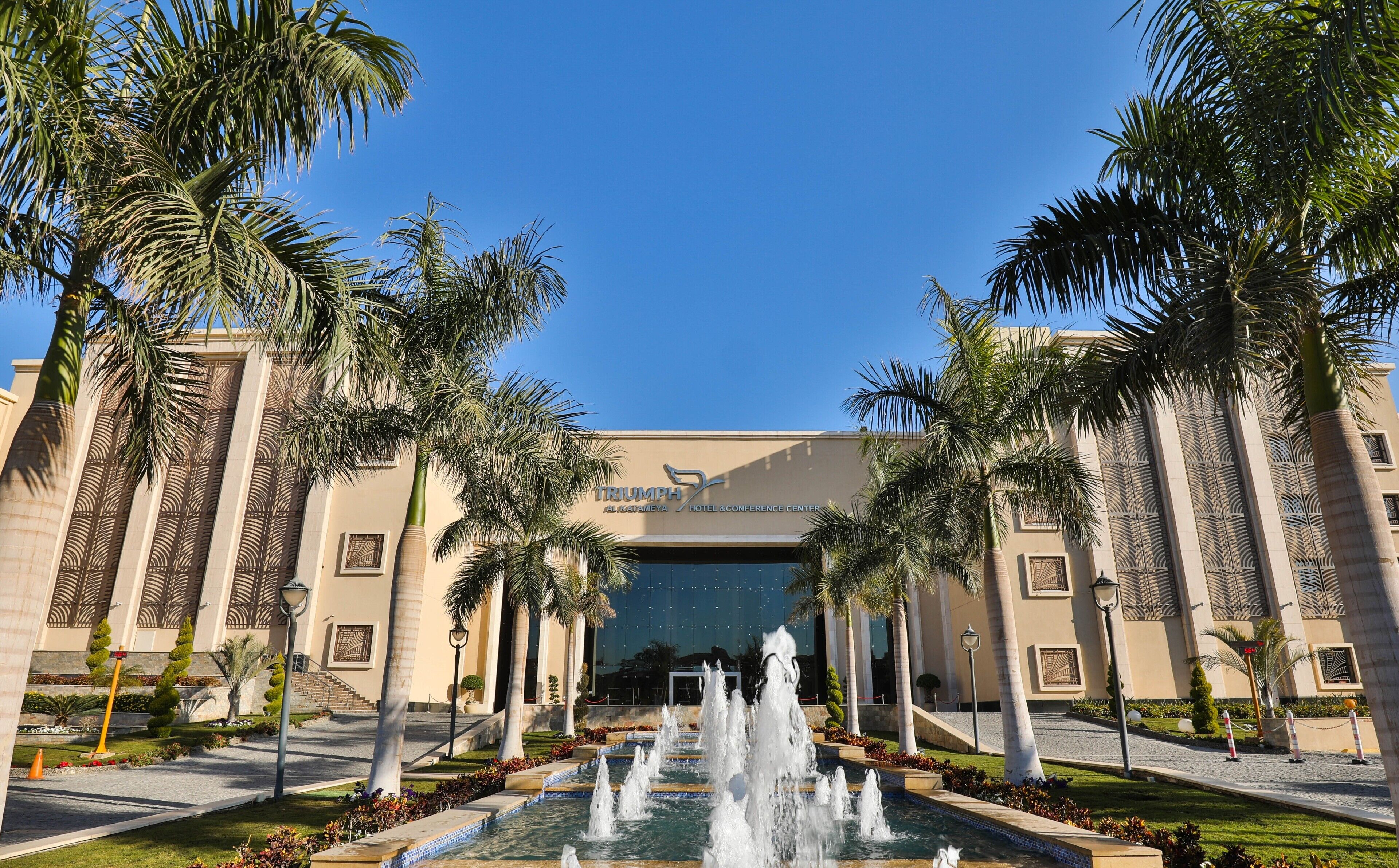 The exterior of the Triumph Hotel & Conference Center with a central water fountain, palm trees, and a clear blue sky.