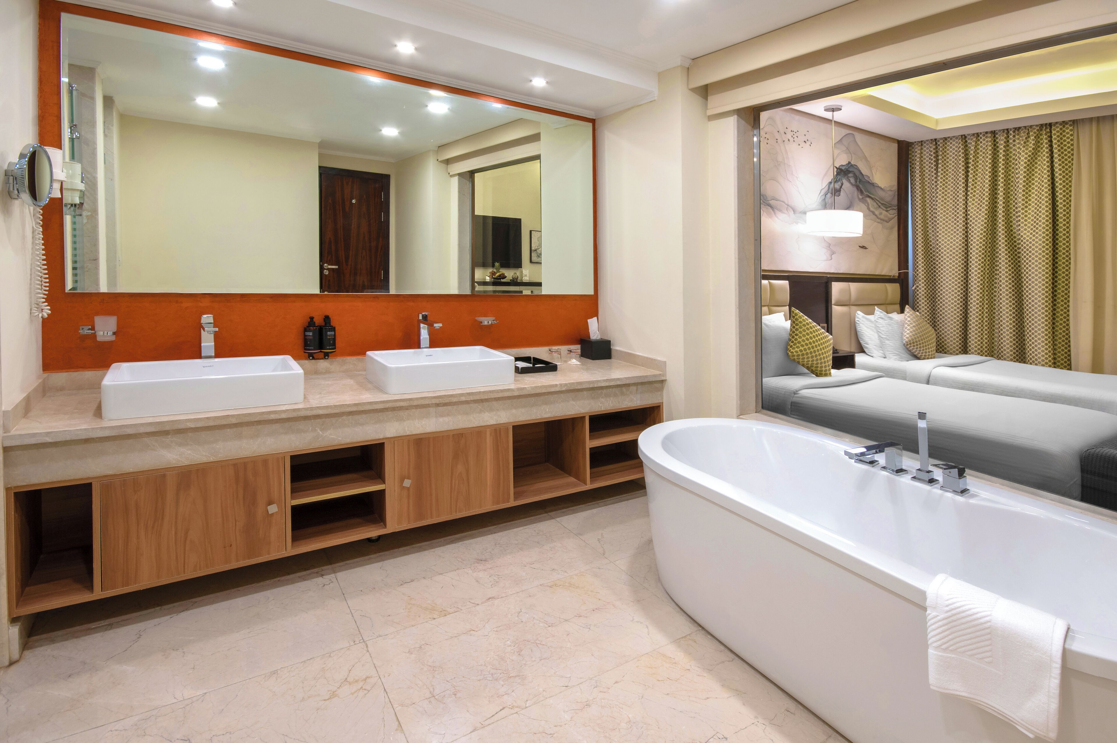 A modern bathroom with two sinks, a large mirror, a bathtub, and a bed visible through an open door.