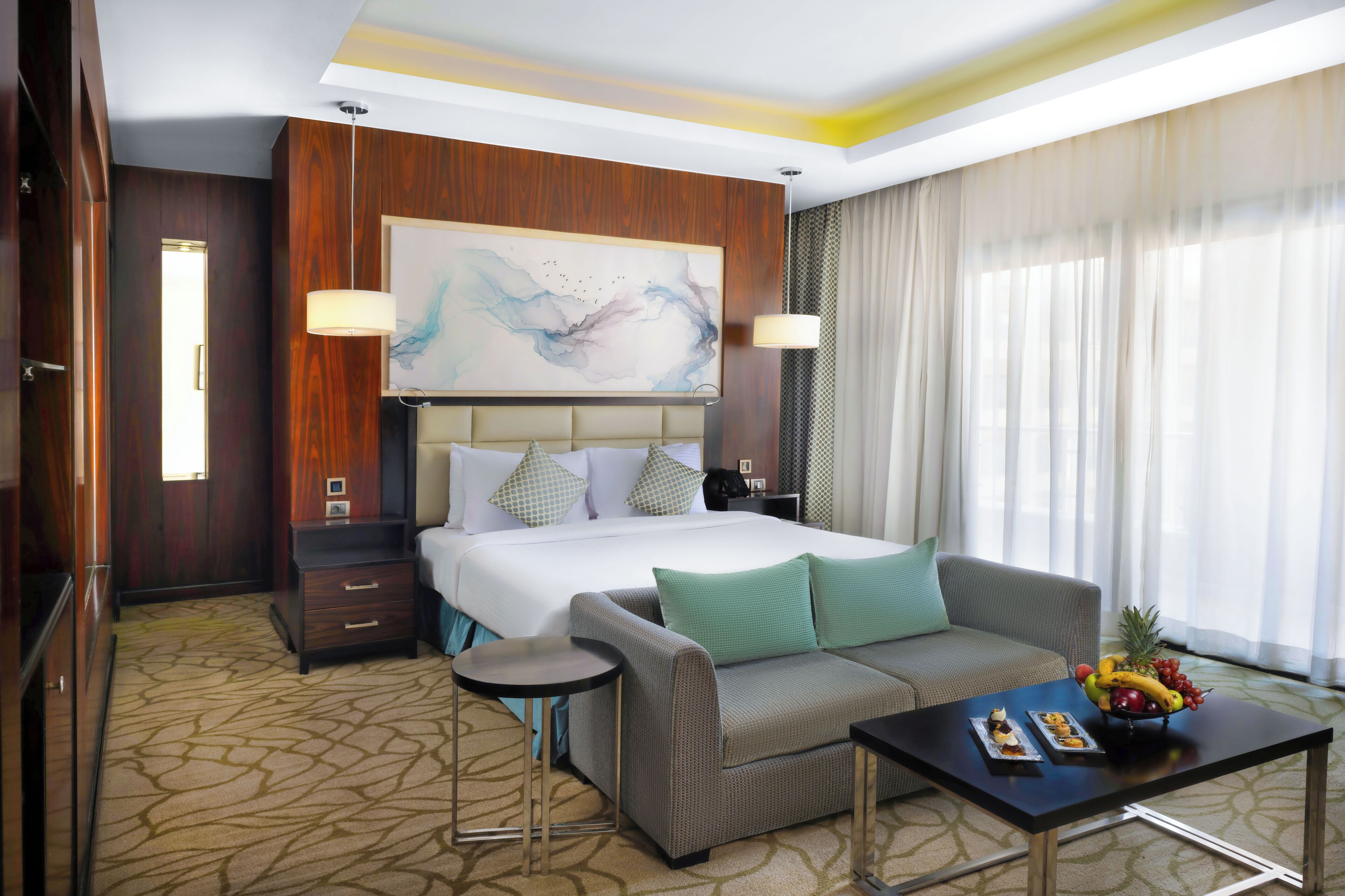 A hotel room with a large bed, a sofa, a coffee table with fruit, and a painting on the wall.