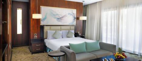 A hotel room with a large bed, a sofa, a coffee table with fruit, and a painting on the wall.