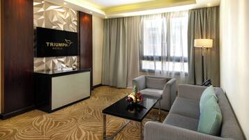 A hotel room with a flat-screen TV, a reception desk, a sofa, a coffee table with fruit, and patterned curtains.