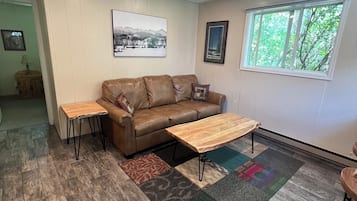 A living room with a brown leather sofa, two wooden side tables, a patterned rug, and a window with a view of trees.