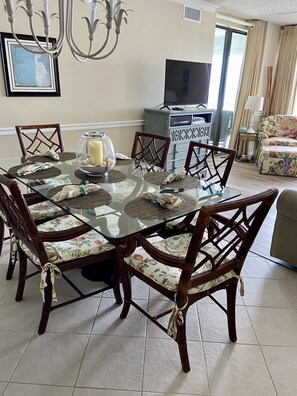 Un comedor con mesa de vidrio, sillas con cojines estampados, una lámpara de araña, un televisor y un sofá floral.