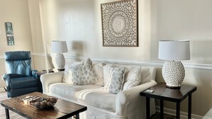 Una sala de estar con un sofá blanco, dos mesas auxiliares con lámparas, una mesa de centro, un sillón de cuero azul y un elemento decorativo en la pared.