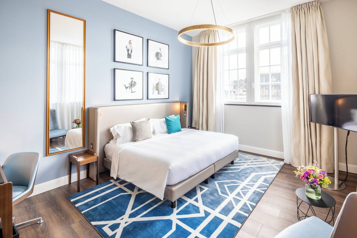 A modern hotel room with a large bed, a bedside table, a chair, a desk, a large mirror, and a window with curtains.