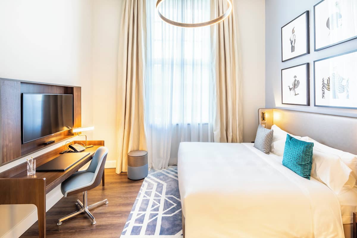A hotel room with a bed, a desk with a TV, a chair, and a window with curtains.