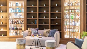 A bar area with a sofa, ottomans, and a round table with drinks. Shelves with bottles are visible in the background. - Fraser Suites Hamburg (Hamburg)