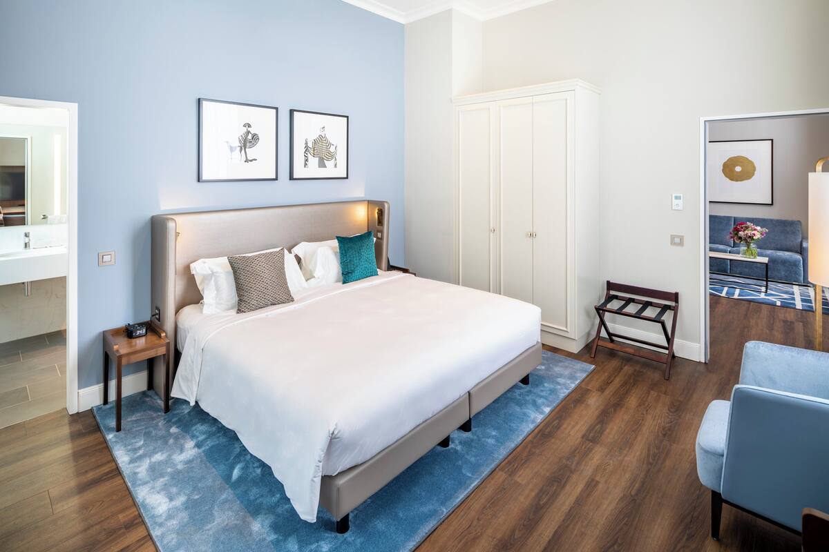 A bedroom with a large bed, a nightstand, a dresser, a chair, and a wardrobe. There are two framed pictures on the wall.