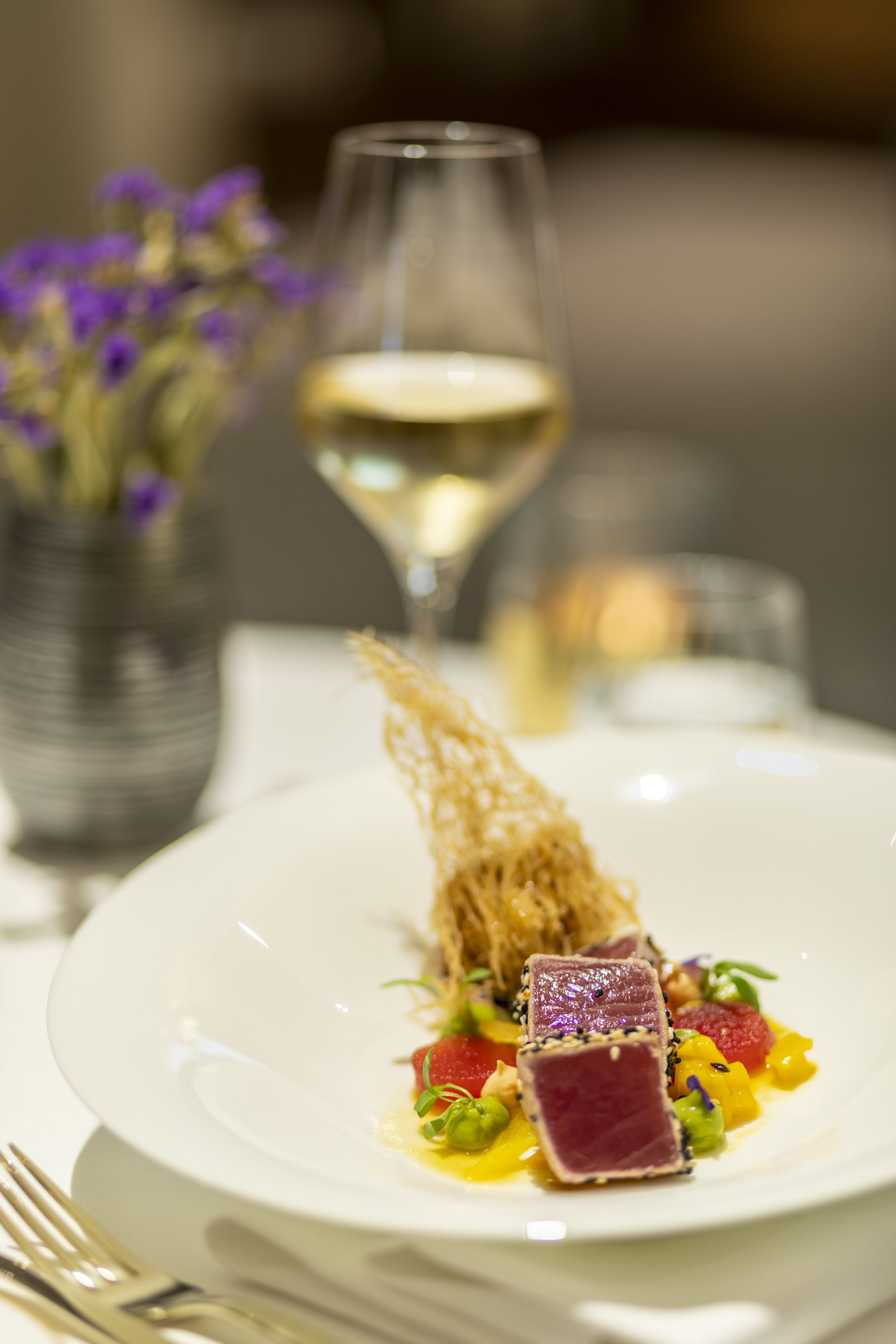 A white plate with a dish of food, a glass of white wine, and a candle in the background.