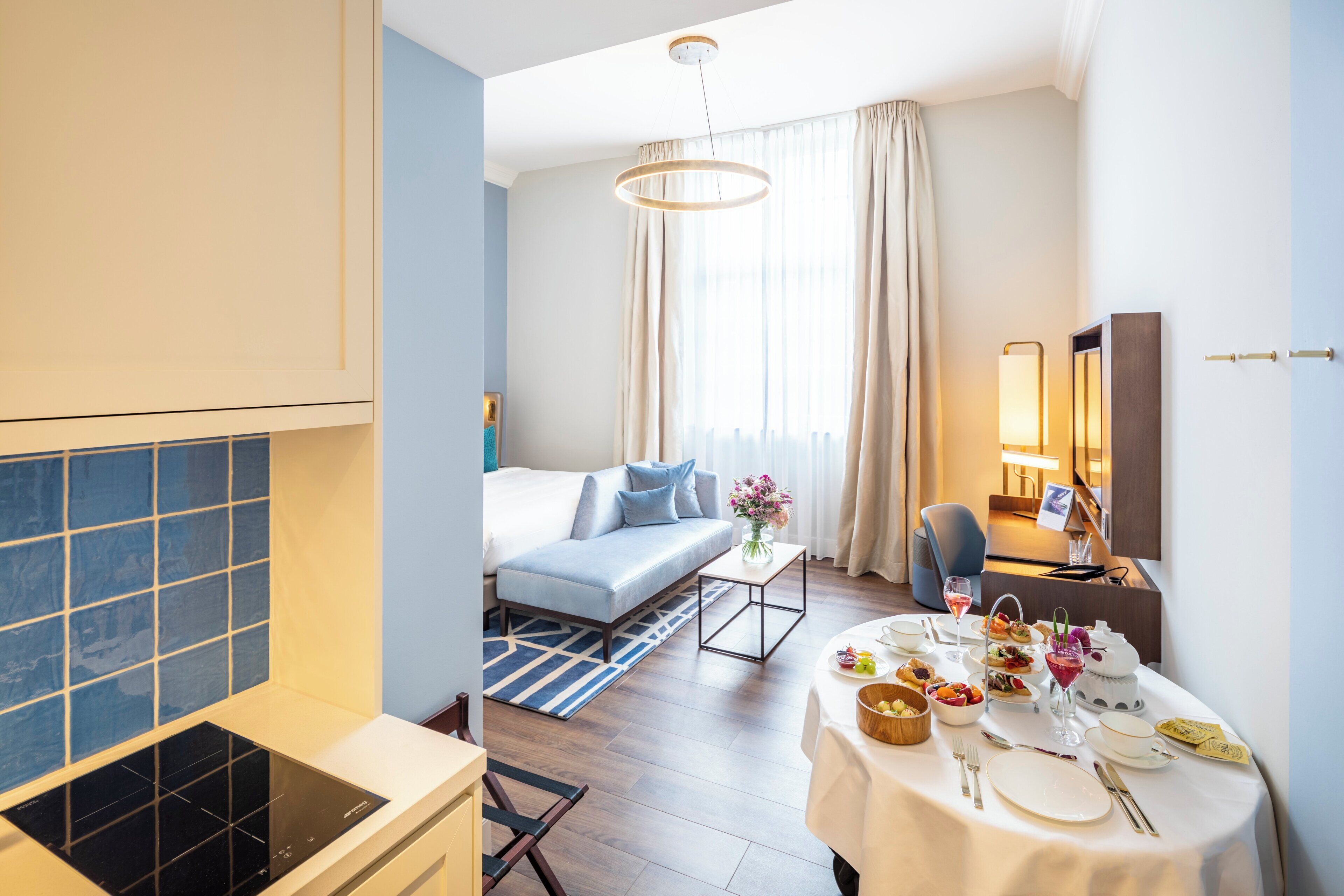 A hotel room with a sofa, dining table set for a meal, a small kitchen area, and a large window with curtains.