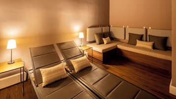 A modern hotel room with a sofa, two reclining chairs, a small table, and a lamp.