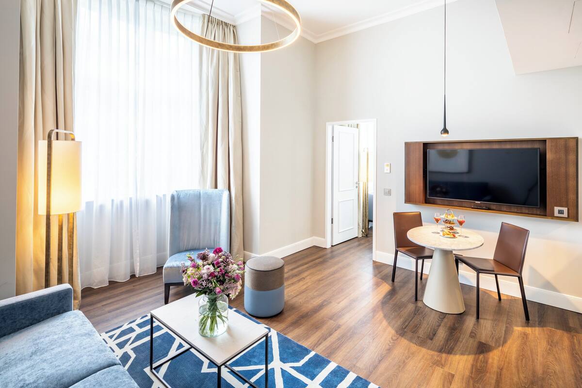 A modern hotel room with a living area, a dining table, and a TV mounted on the wall.