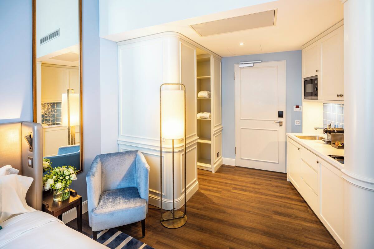 A hotel room with a bed, a chair, a floor lamp, a vanity with a mirror, a microwave, and a door leading to a bathroom.