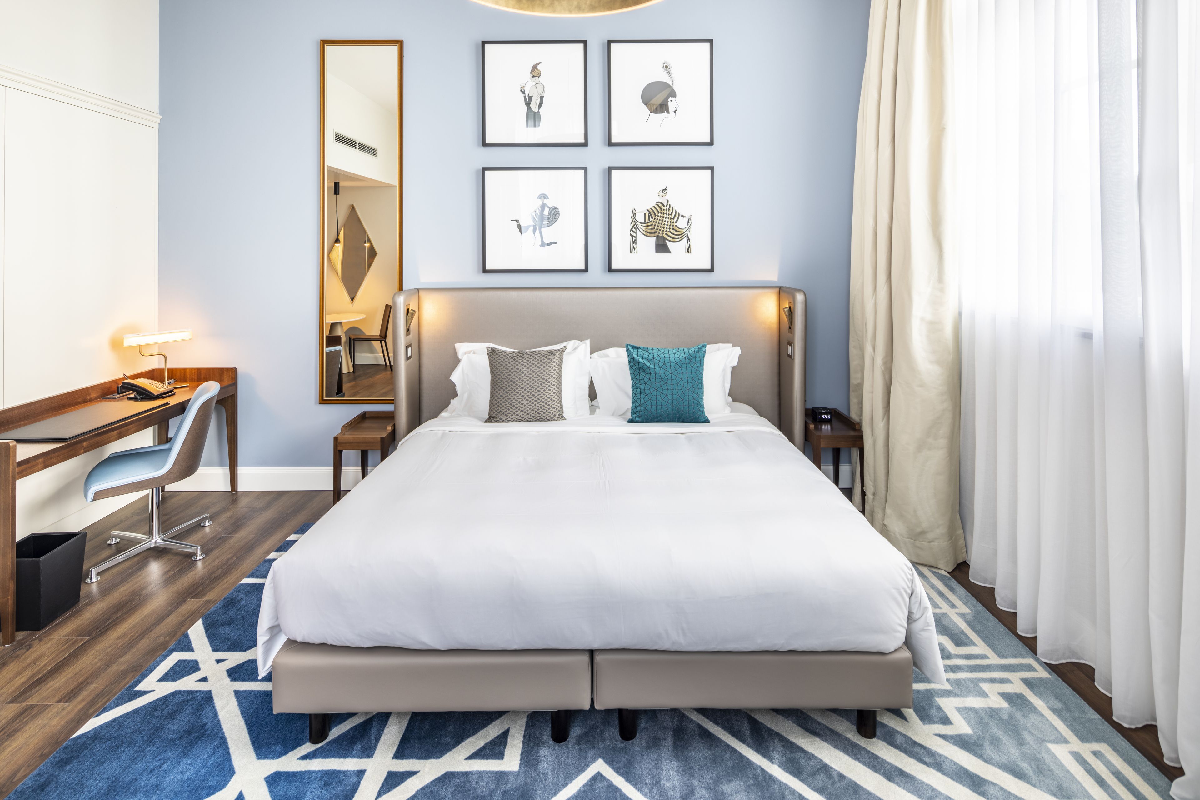 A hotel room with a large bed, a desk, a chair, and a mirror. There are four framed pictures on the wall.