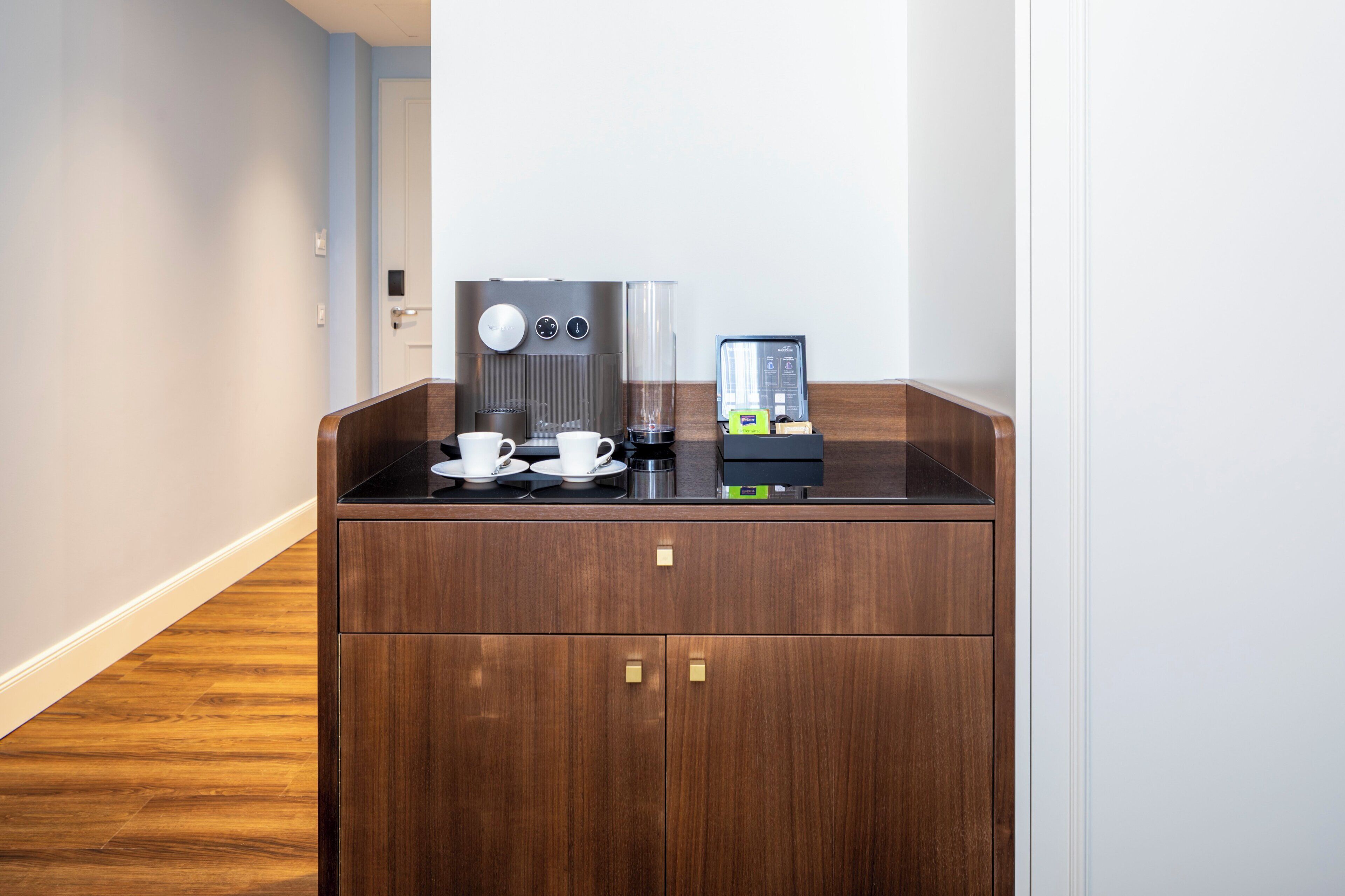Estação de café com cafeteira, xícaras e dispensador de cápsulas em um quarto de hotel.