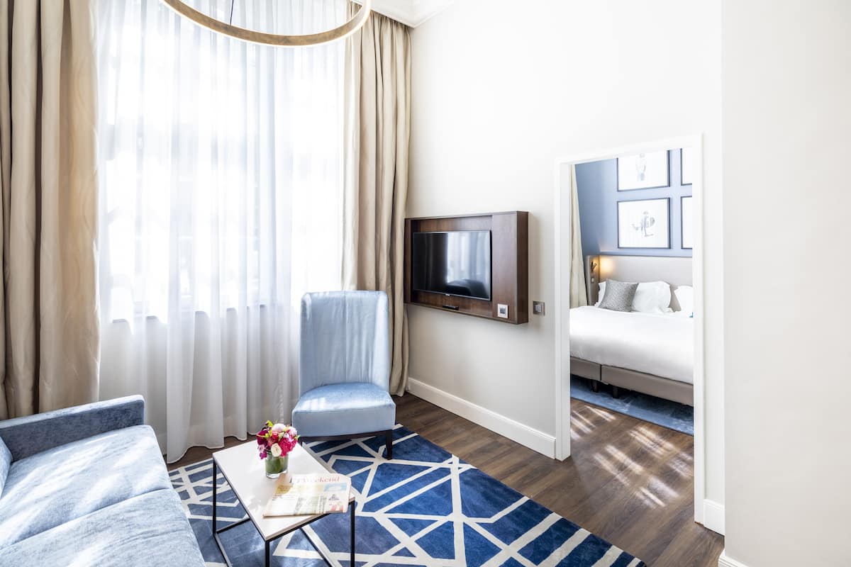 A modern hotel room with a large window, a TV mounted on the wall, a sofa, a chair, and a bed.