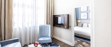 A modern hotel room with a large window, a TV mounted on the wall, a sofa, a chair, and a bed.