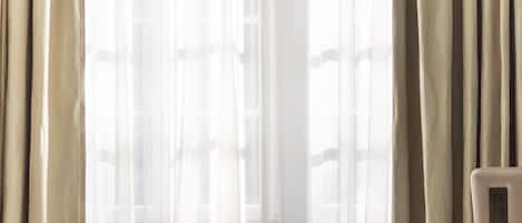Una habitación de hotel con una cama grande, cortinas color beige y un ventanal con cortinas traslúcidas.
