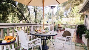 Une terrasse couverte avec une table dressée pour un repas, entourée de chaises en osier blanc, et une vue sur une zone boisée.