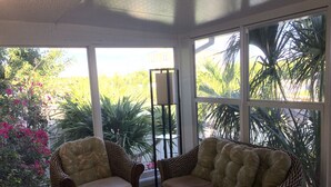 Une véranda meublée de mobilier en osier, dotée de grandes fenêtres et agrémentée de plantes tropicales.