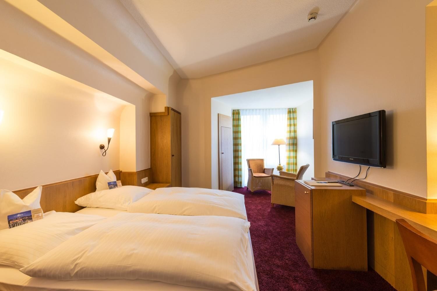 Una camera d'albergo con due letti, una scrivania, una sedia, una televisione e una finestra con tende.