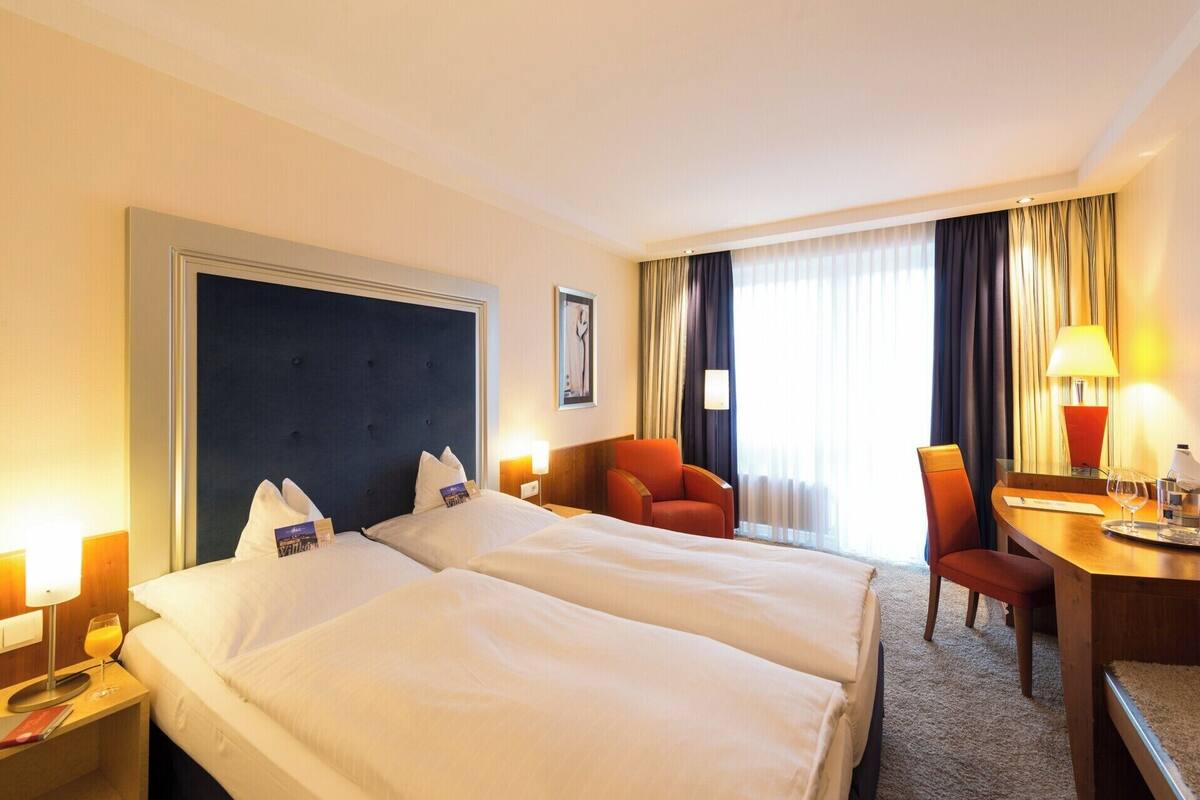 Habitación de hotel con dos camas, un escritorio, una silla y un ventanal con cortinas.