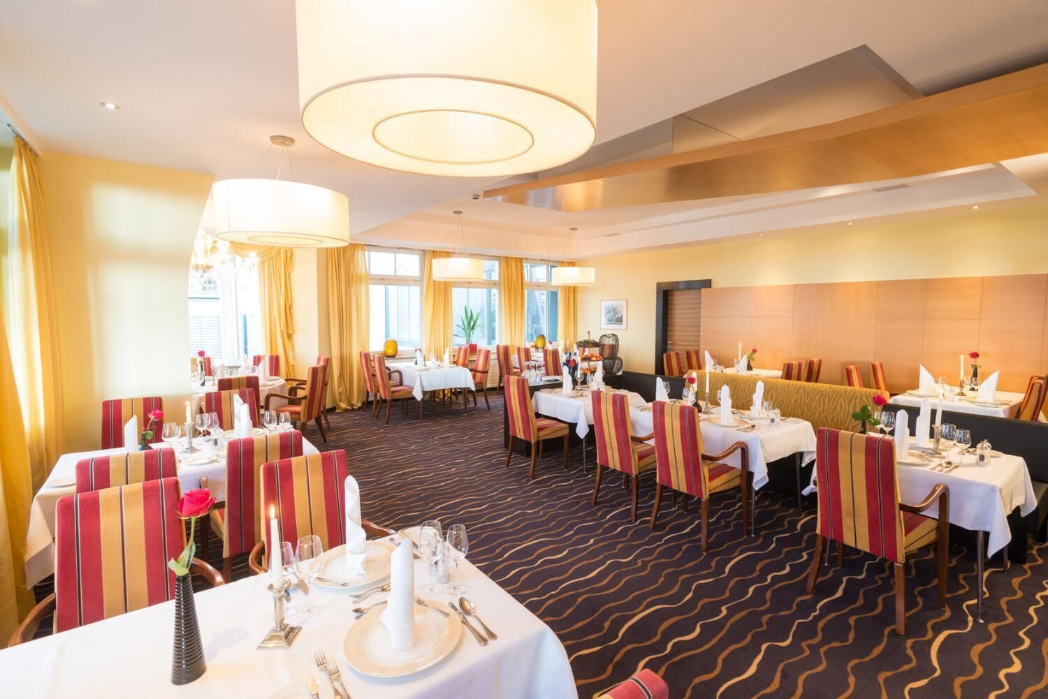 A hotel dining area with multiple tables set for dining, striped chairs, and a large chandelier.