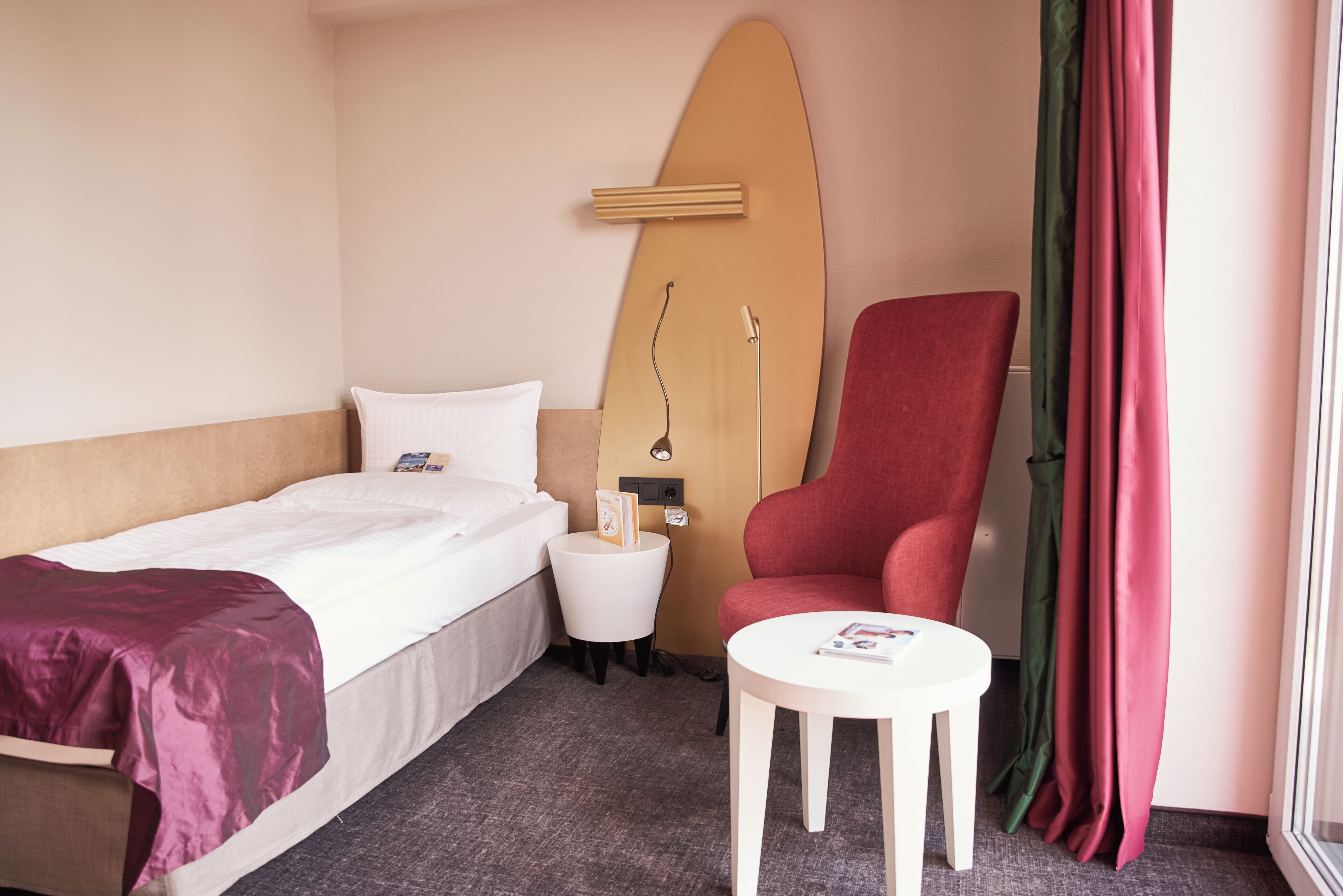 A hotel room with a bed, a red armchair, a small table, and a surfboard mounted on the wall.