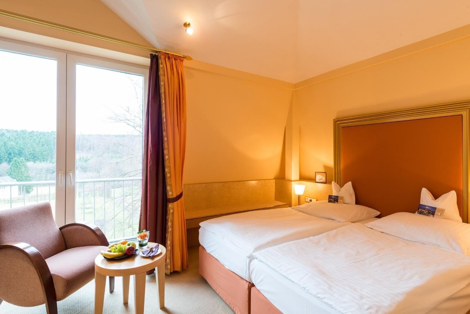 Habitación de hotel con dos camas, una silla, una mesita con fruta y vistas a árboles a través de la ventana.